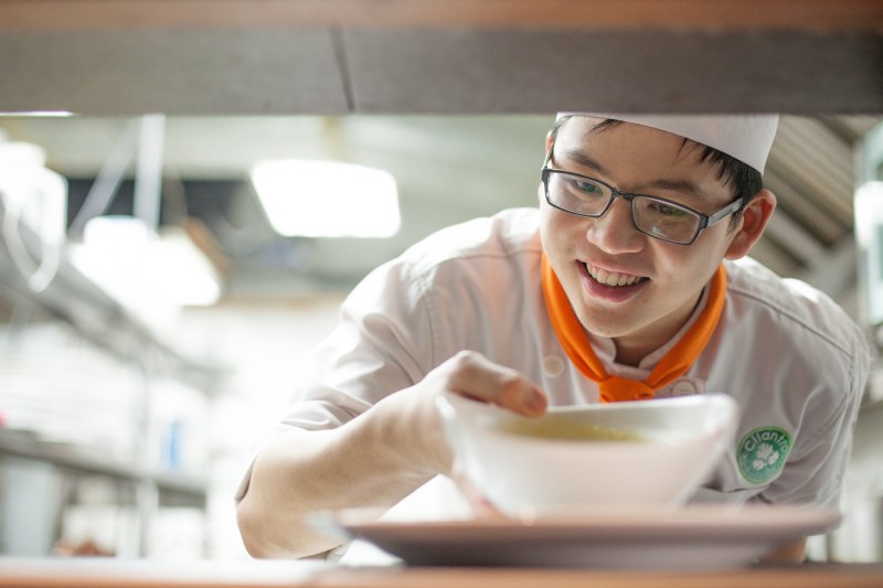 学生面带灿烂的笑容，熟练地准备一道菜。他对烹饪的热情展露无遗，细心地组合食材，加入自己独特的创意